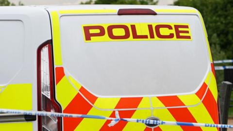 The back of a van with orange and yellow high vis stripes on it and police written in red across the rear door. Blue and white police cordon tape is stretched in front of the van and behind it.