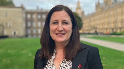 Julia has brown shoulder length hair and is smiling. On her right is the houses of parliament which are large brown buildings. She is wearing a black suit jacket and black and white blouse, with a red rose pin on the jacket