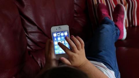 A child turned away from the camera wearing a tshirt, jeans and red trainers, lies back on a brown sofa while using a mobile phone