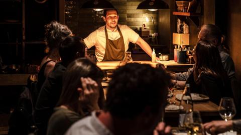 Chef Corrin in kitchen which is open to dining area talking to diners at two tables together. The setting is dark with warm overhead lighting and the diners are all looking at him.