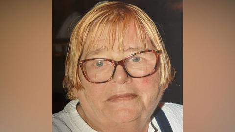 Delia Sheridan stares at the camera. She has short blonde hair and glasses. She is wearing a white top.