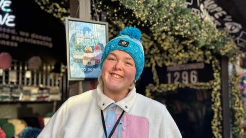 A girl wearing a blue hat and white top with a pink pocket is smiling to the camera. She is front of a Christmas market stall which sells hats.