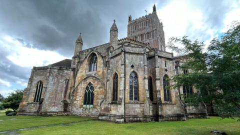 A view of Tewkesbury Abbey - a grand old church building with trees next to it, and a grass lawn.