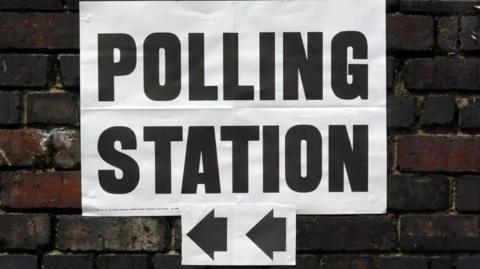 A white polling station sign with black letters and arrows pointing to the left. The sign is on a brick wall