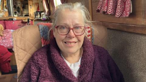 A woman with grey hair, red-framed glasses and a maroon jumper in a brown chair sitting on board her narrowboat. A small room can be seen behind her.