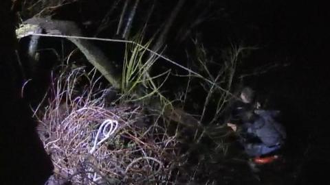 Two men cling to a broken tree branch which is dangling into the river. It is night. a rope stretches over the branch from the river brank down to the teenagers.