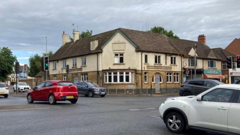 White and brown pub building on a corner next to a road junction with cars going past