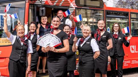 A group of eight women and one man wearing Loganair uniforms smiling at the camera. They are standing in front of a red bus and are waving French flags. The steward at the front is holding a bunch of flight tickets fanned out. The unform is black and the women are wearing red checked scarfs.
