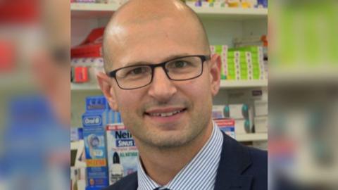 Olivier Picard smiling at the camera, wearing dark framed glasses and a blue stripe shirt with navy blazer.