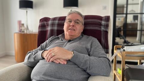 Steven Barrett sits in an armchair in a living room, facing the camera and smiling. He is wearing a grey fleece and glasses.