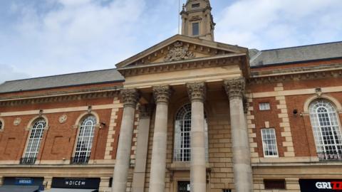 The Peterborough City Council building’s exterior combines red brick with light-colored stone detailing around the windows and columns. Large arched windows line the front, and at street level, modern shop signs such as “Greggs,” “The Dice Bar,” and another partially visible sign are present, indicating the building now houses commercial businesses. The sky above is partly cloudy, adding a soft contrast to the architectural details.