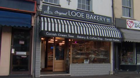 Looe Bakery with a sign saying its name in white on a black background. It has a black and white pattern of stripes above it and also says Cornish Cream Teas, Bakery & Coffee Shop and Cornish Pasties.