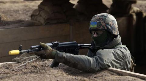 A Ukrainian military recruit takes part in a training exercise conducted by British and Dutch army personnel in the north of England in February 2023. The recruit is in a trench holding a rifle. He is wearing camouflage and a helmet with the Ukrainian flag on it. 