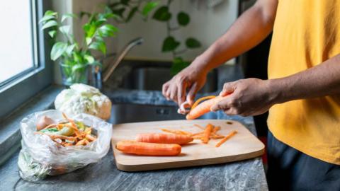 A person peels carrots over a wooden chopping board. They have a bag next to them with the peelings in and with other food waste. In the background there is a sink and plants.