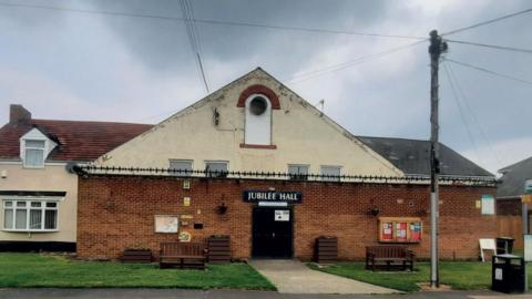 Jubilee Hall in West Rainton. The extension is made of brown bricks and has sharp anti-theft spikes running along its roof. The yellow paint is flaking off the roof and a window has been bricked up.