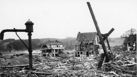 A black and white photograph showing old-fashioned rural German buildings, with timber frames, destroyed in the Dambusters raid, there is debris piled up around the remains of the buildings. 