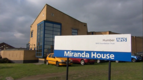 The outside of Miranda House, Hull. The yellow brick building has blue window frames and stands behind a blue and white NHS sign.