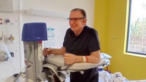 A man with black, grey hair stands with the help of a medical support in a hospital room which has a white wall and a yellow wall behind him. He wears half-rimmed glasses and a blue top.