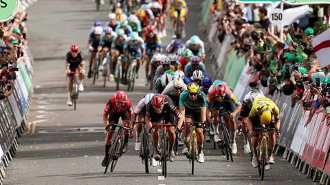 A group of cyclists racing against each other on a road. Members of the public are gathered behind barriers watching.