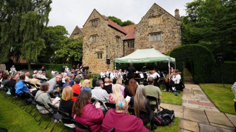 Dozens of people sit on chairs in the garden of the tall stone building. There are watching a brass band playing under a green National Trust gazebo. There are tall, lush green trees in the background.