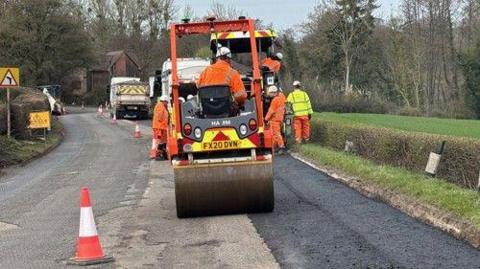 Workmen resurfacing a country lane, they are wearing orange and yellow hi-vis. 