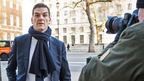 Olly Robbins wearing a suit and scarf and walking through london. The arm and partial face of a photograher can be seen pointing a camera at him 