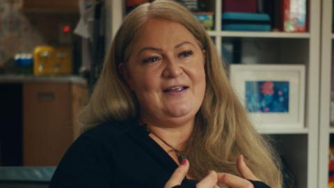 Lynn McDonald with long, blonde hair sits on a sofa. She wears a black top. There is a bookcase behind her with books and photoframes.