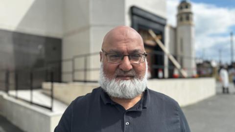 Zaf Iqbal, wearing glasses and a black t-shirt, standing outside the Masjid E Anwaar E Madinah mosque in Sunderland