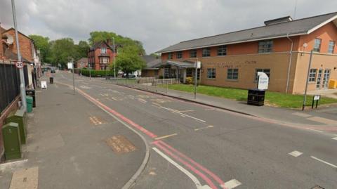 A Google street view image of a road in the middle of a residential area. A community centre is on the right of the road. A 20mph sign is on a post on the left. 