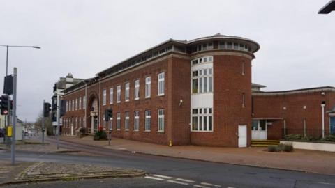 Torquay police station. It is a large red brick building with dozens of windows next to a road with traffic lights on it. It is an overcast day.
