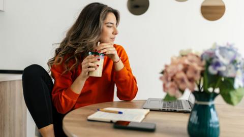 Woman with long, light brown hair wearing an orange long-sleeved top and black leggings sits at a table at home drinking a smoothie and looking at a laptop