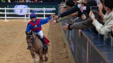 Harriet dressed in a blue and red riding costume riding her light brown Shetland Pony, holding her arm out for the crowd who are reaching towards her for a  high five
