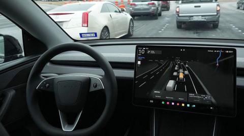 A black steering wheel and large screen to the right which shows lanes of traffic and vehicles in it.
