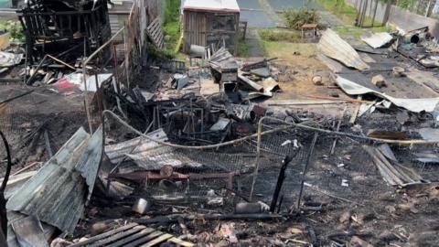 A picture of the charred remains of the allotments. There is twisted metal, burnt wood and what remains of the chicken coop. There is a shed in the background.