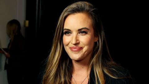 Charlie Webster smiles at the camera. She has long brow hair and is wearing a black jacket and top.