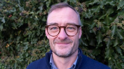 Tom Surrey wearing glasses and smiling at the camera. He has short hair and a short beard. He is wearing a blue jumper with a blue shirt underneath. He is stood in front of tall green bush with small flowers.