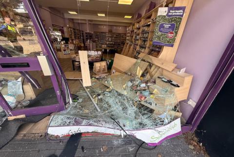 The picture shows the front of a store that has been destroyed. There is broken glass on the floor, the doors are bent inwards, shelves have fallen along with the goods in them. The back of the store is completely untouched, as products can be seen lined up neatly on shelves.