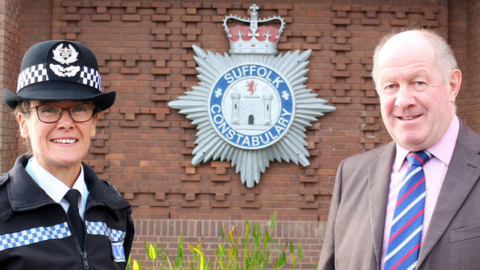 A woman wearing a police officer's uniform next to a bald man wearing a suit with a pink shirt and a multi-coloured tie. They are standing in front of a brick wall with the Suffolk Police emblem on it. 