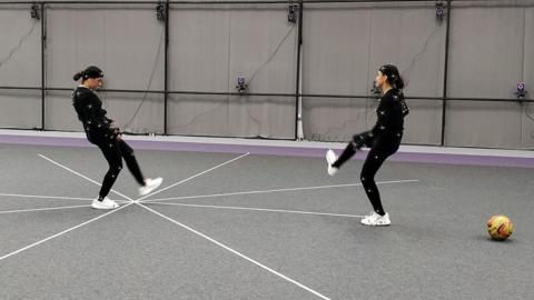 Two women in skin-tight black motion-capture suits dotted with small ping-pong style balls make kicking motions inside a studio. Cameras are placed at regular intervals on the grey walls and white tape in a star arrangment marks reference points on the floor. 