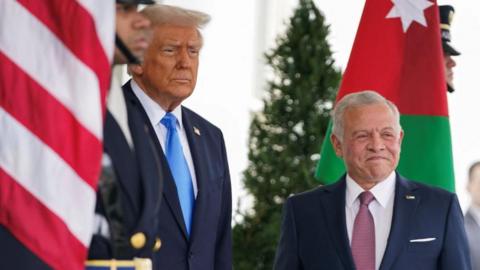 US President Donald Trump meets with Jordan's King Abdullah at the White House in Washington. The king is smiling and you can see both the US and Jordanian flags. 