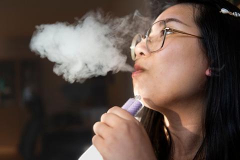 A young person exhaling smoke from a vape, which she is holding in her hand. 