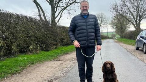 Councillor Paul Hopton - a man with short grey hair smiling with a navy coat standing along a single-access lane with his dog sitting by his side on a lead. The dog is a brown cocker spaniel.