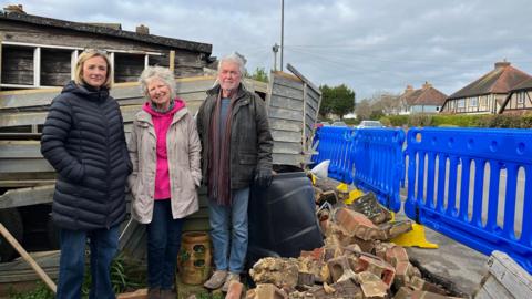 Karen Vilday, left, Sue and Des Mills standing in Karen's back garden where the wall has been destroyed by a crashed car. The bricks are scattered on the floor and blue plastic fencing has been put up on the pavement next to the road which is a metre away. Wooden fencing has also been destroyed. All three are wearing winter jackets and looking at the camera. 