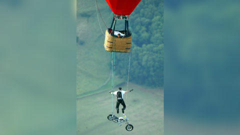 A man leaps from a motorcycle suspended below a hot air balloon