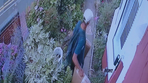 A man walks away from the front door of a house with two brown parcels in his hands. He has a cap on and bad on his back as he walks on a short garden path through a front garden away form a house.