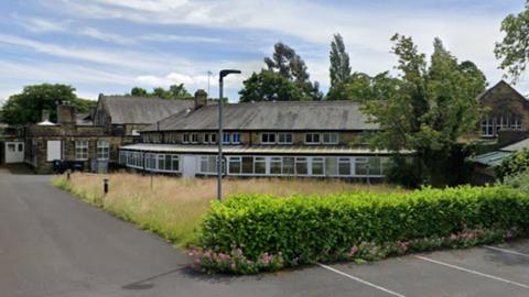 A large two storey stone building with a large overgrown garden and car park in front