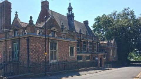 The ornate brick building has a turret with what looks like a bell tower.  There are lots of stone pinnacles on the roof.  The building is surrounded by railings and a tree grows next to it on the right.