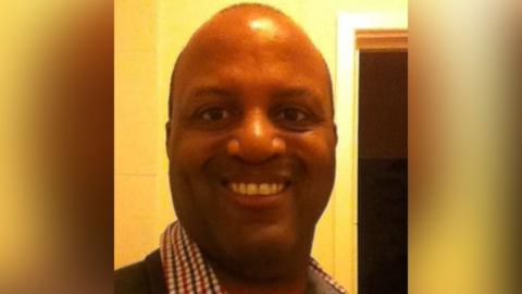 A head and shoulders selfie image of a man smiling at the camera, wearing a red white and blue checked shirt and dark jumper. He has brown eyes and has thinning dark hair