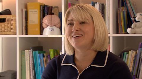 Dr Sarah Mills is wearing a navy blue collared top with white edging and has a blonde bob and fringe. She's standing in front of a bookshelf containing assorted books and files, along with some ornaments which include what looks like a mini model brain.
