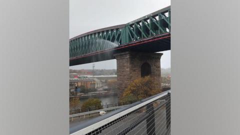 The Queen Alexandra Bridge. The green steel bridge has a water pipe running along its side. Water is spraying from the pipe onto the River Wear below.